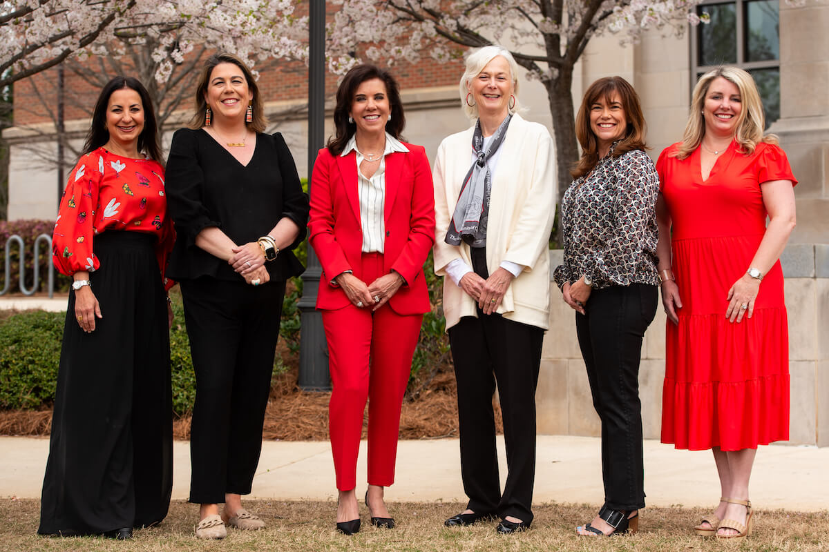 EC-2024-GWGWebUpdates-V1 Six women pose together outdoors under blooming trees near a brick building.