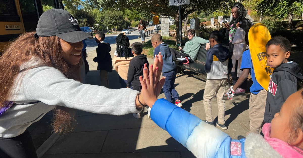 UGA Ambassador high-fiving a young student among a larger group of students near school bus. UGA Ambassador high-fiving a young student among a larger group of students near school bus.