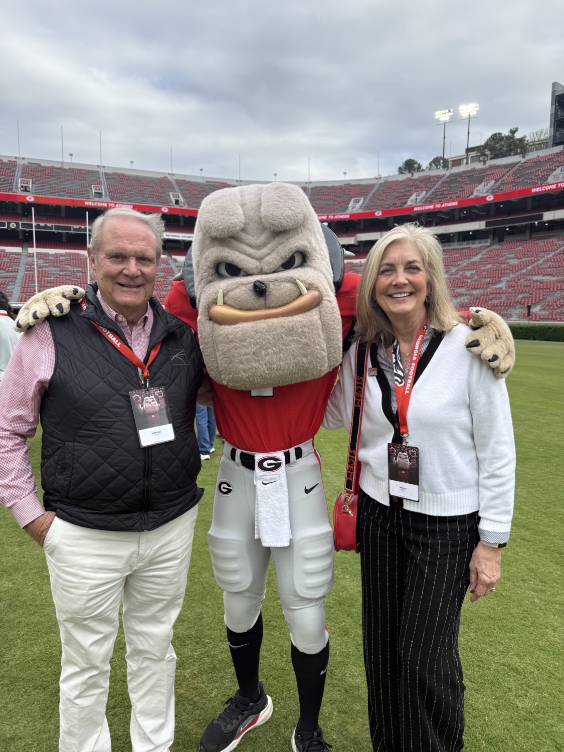 A smiling man and woman stand on either side of a dog mascot on a football field.