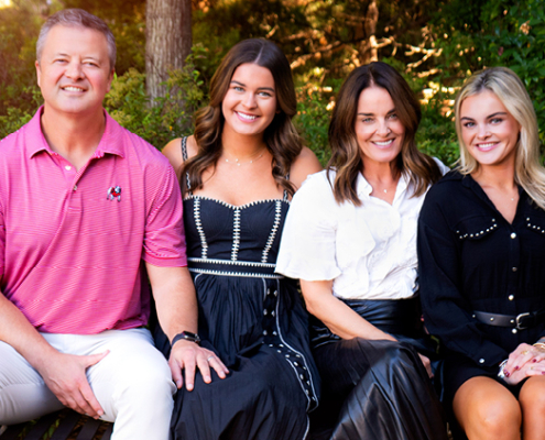 A father, mother and two daughters sit on a bench on a college campus on a sunny afternoon.